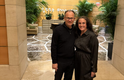Two people stand together and smile in a lobby with marble floors, green plants, and a wooden wall with white orchids in the background.