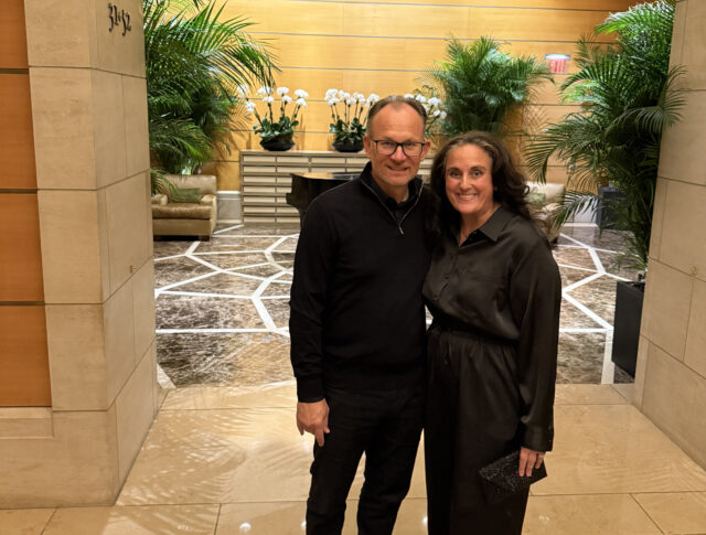 Two people stand together and smile in a lobby with marble floors, green plants, and a wooden wall with white orchids in the background.