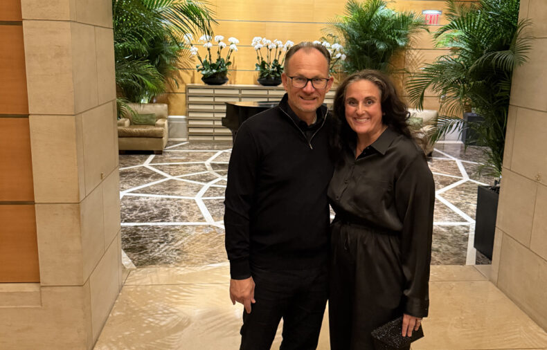 Two people stand together and smile in a lobby with marble floors, green plants, and a wooden wall with white orchids in the background.