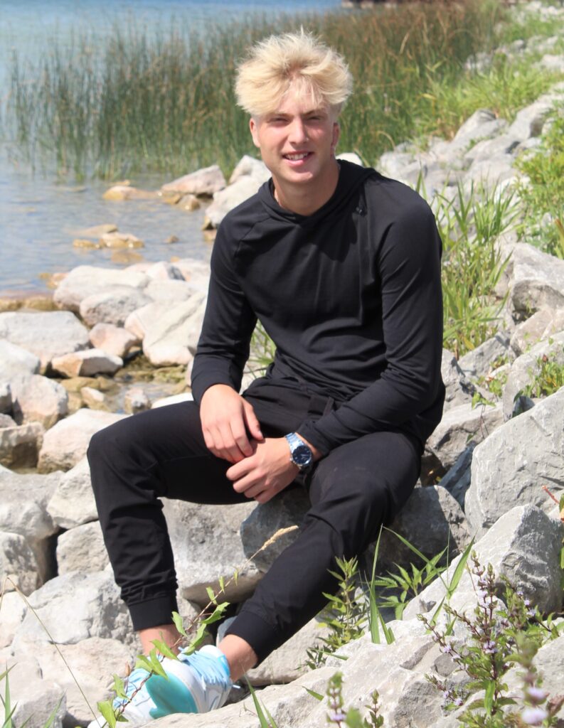 A young man with blond hair, wearing a black tracksuit and blue-green sneakers, sits on rocks near a lake with reeds and grass in the background, reflecting on the immediate impact of his endowed scholarship.