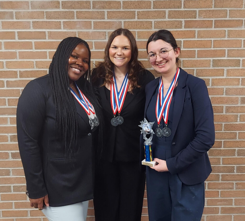 Three women wearing medals stand in front of a brick wall; one holds a small trophy. They are dressed professionally and smiling at the camera.