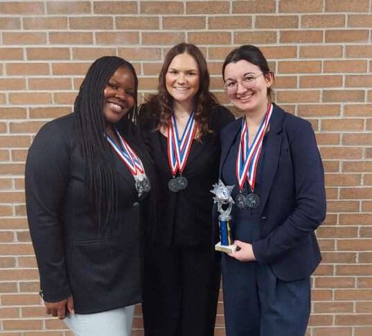Three women wearing medals stand in front of a brick wall; one holds a small trophy. They are dressed professionally and smiling at the camera.