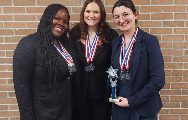Three women wearing medals stand in front of a brick wall; one holds a small trophy. They are dressed professionally and smiling at the camera.