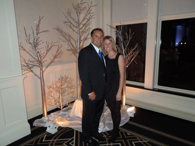 A man in a suit and a woman in a black outfit stand smiling together in front of decorative, leafless trees indoors, celebrating the Immediate Impact of an endowed scholarship.