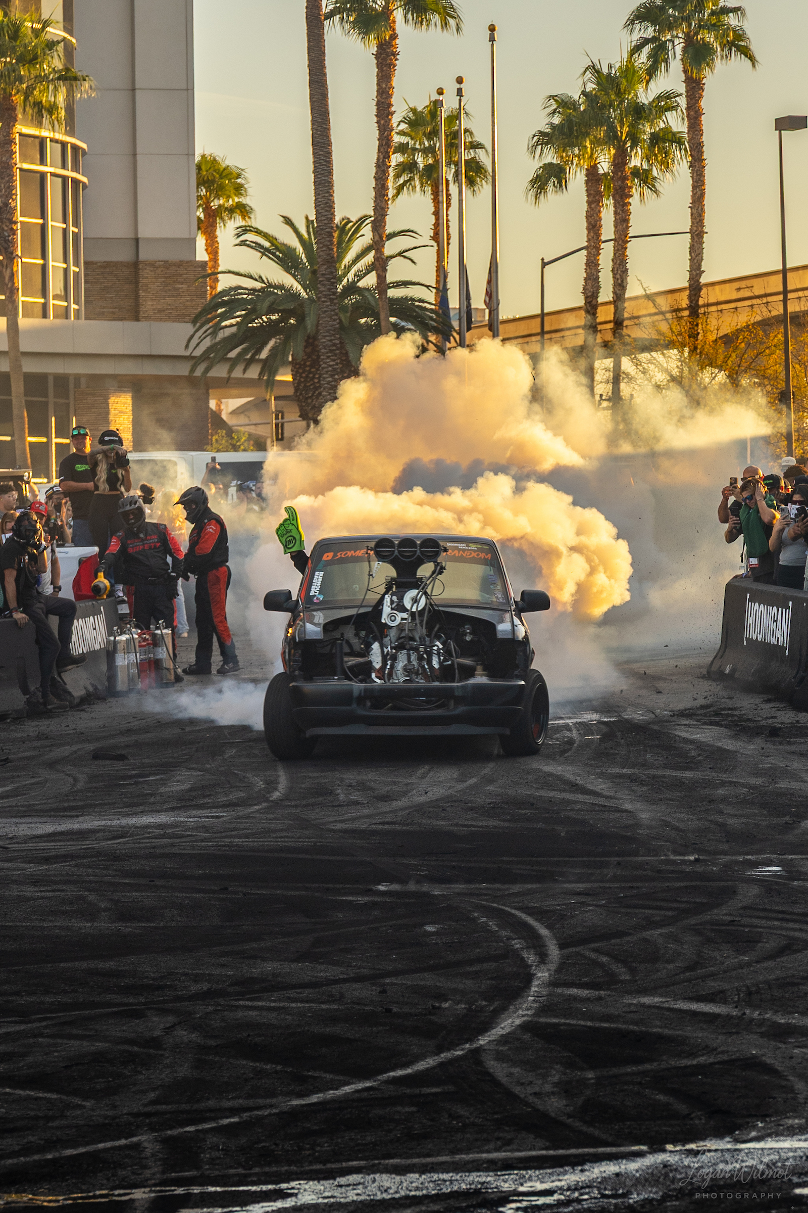 A black car performs a burnout, producing thick white smoke while spectators and photographers watch behind barricades at an outdoor automotive event lined with palm trees.