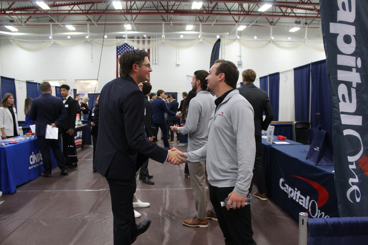Two men shake hands at a career fair, surrounded by booths including Capital One and other companies, with several other people networking in the background—an event often attended by legacy students seeking alumni benefits.