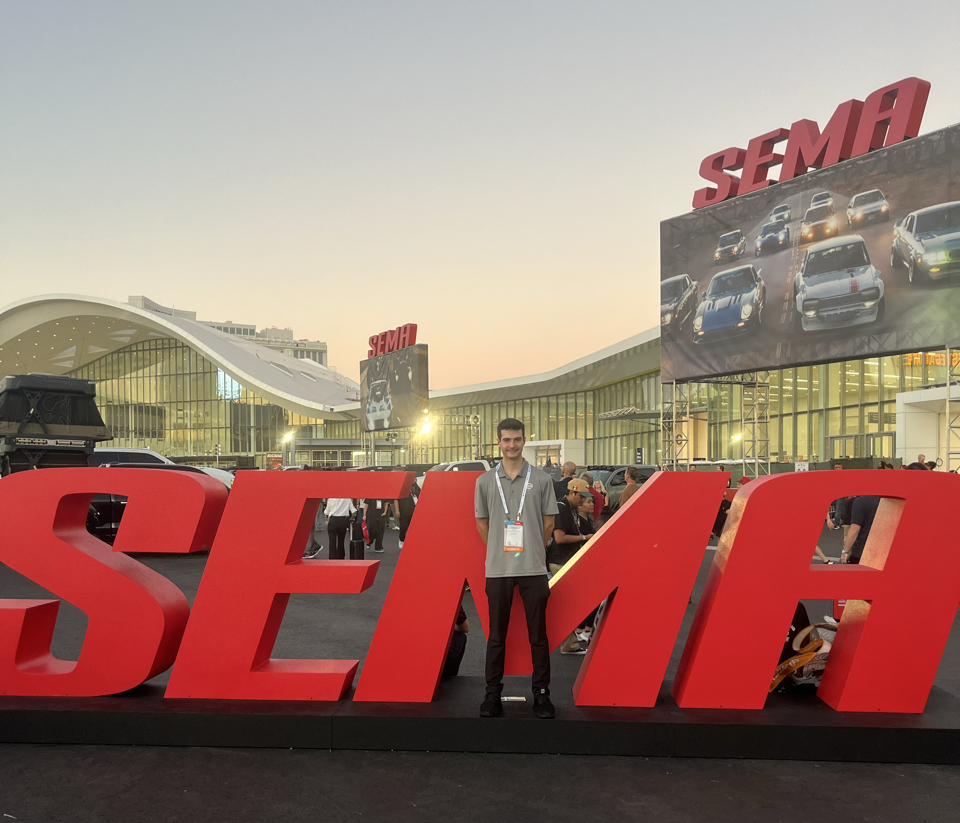 A person stands in front of large red "SEMA" letters outside a convention center at dusk, capturing the excitement of the automotive world with a SEMA sign and car display visible in the background.