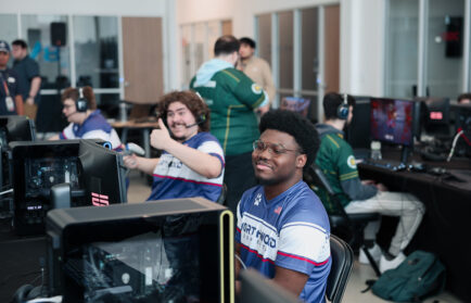Several gamers sit at computer stations in a brightly lit room, with one person smiling at the camera and another giving a thumbs-up gesture.