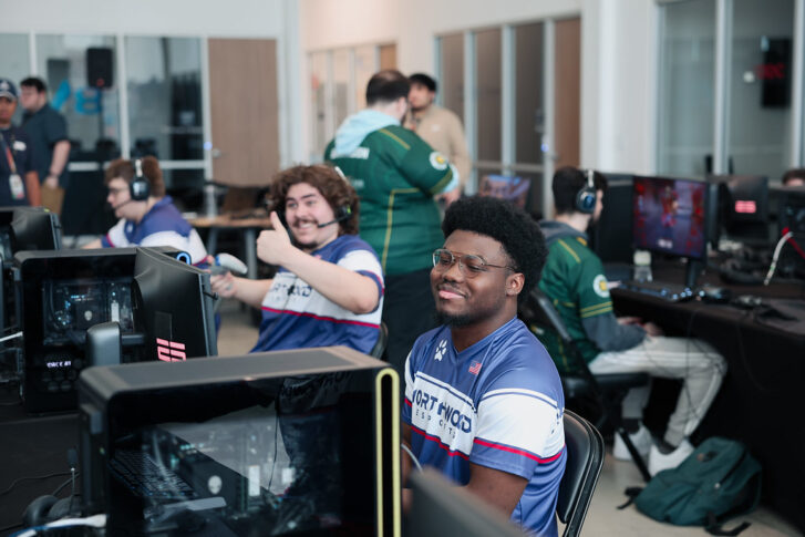 Several gamers sit at computer stations in a brightly lit room, with one person smiling at the camera and another giving a thumbs-up gesture.
