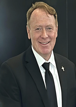 A man in a black suit, white shirt, and black tie with a cross pin on his lapel stands in front of a dark background, smiling at the camera.