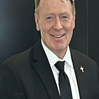 A man in a black suit, white shirt, and black tie with a cross pin on his lapel stands in front of a dark background, smiling at the camera.