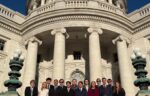 A group of thirteen people in business attire stands in front of a large government building with columns and ornate architectural details.