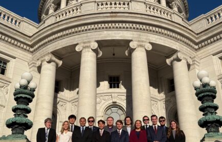 A group of thirteen people in business attire stands in front of a large government building with columns and ornate architectural details.