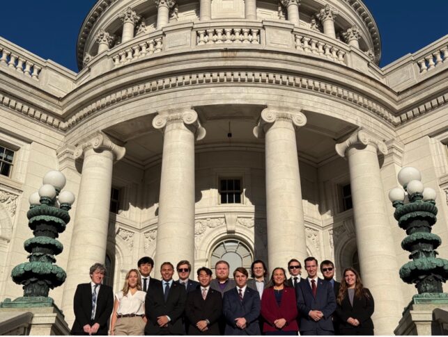 A group of thirteen people in business attire stands in front of a large government building with columns and ornate architectural details.
