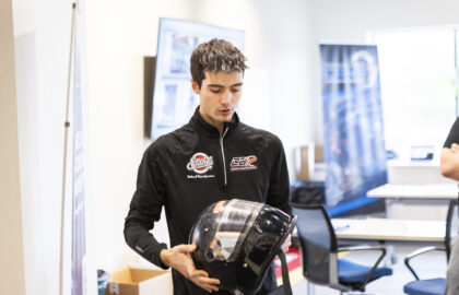 A person in a racing suit holding a black helmet stands in a brightly lit office-like room, suggesting an environment focused on Automotive mobility, with chairs and banners visible in the background.