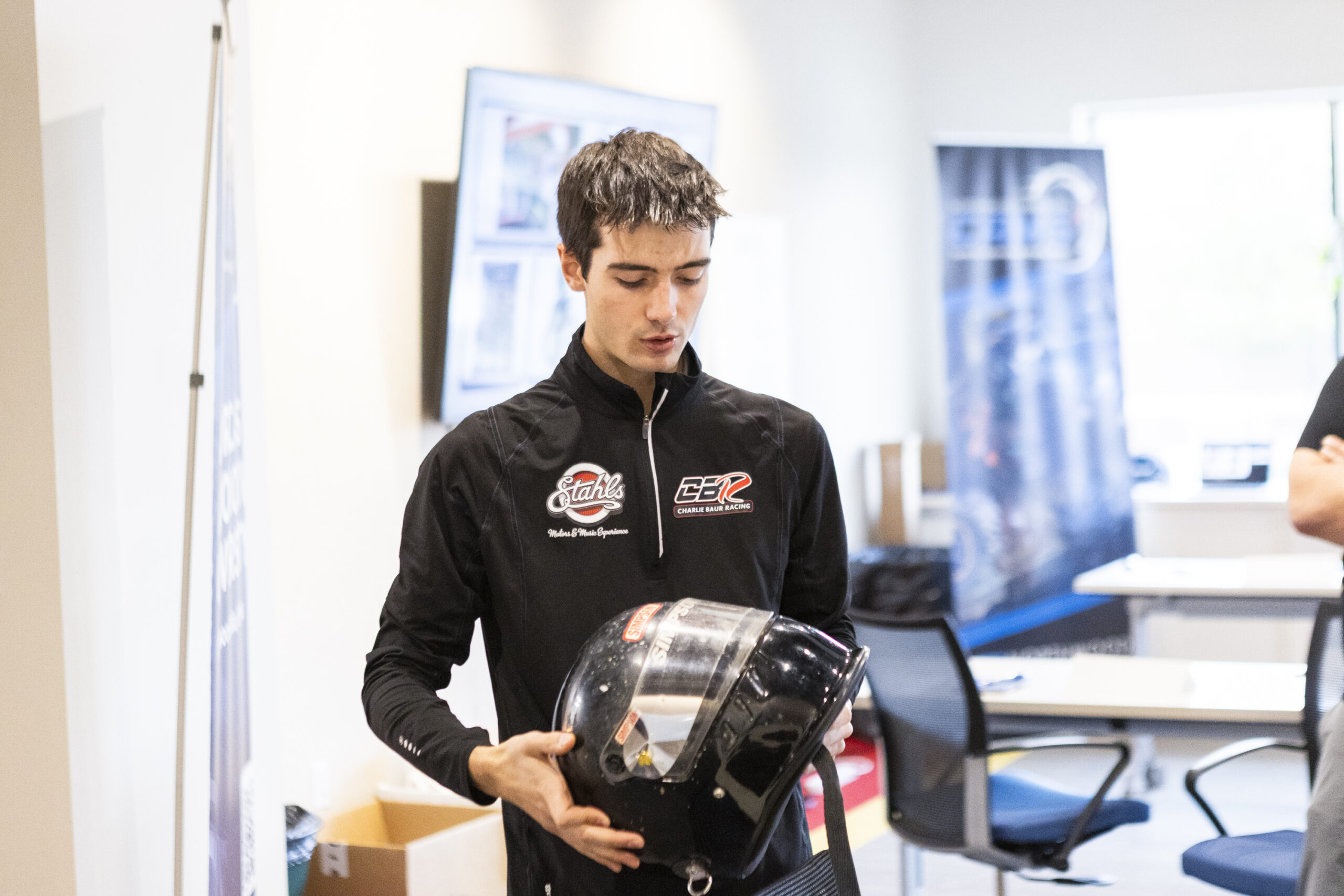 A person in a racing suit holding a black helmet stands in a brightly lit office-like room, suggesting an environment focused on Automotive mobility, with chairs and banners visible in the background.
