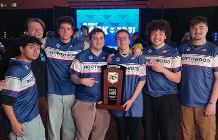 A group of seven young men in matching blue "Northwoods Esports" jerseys pose indoors, holding a large "Champion Fall 2023" trophy.