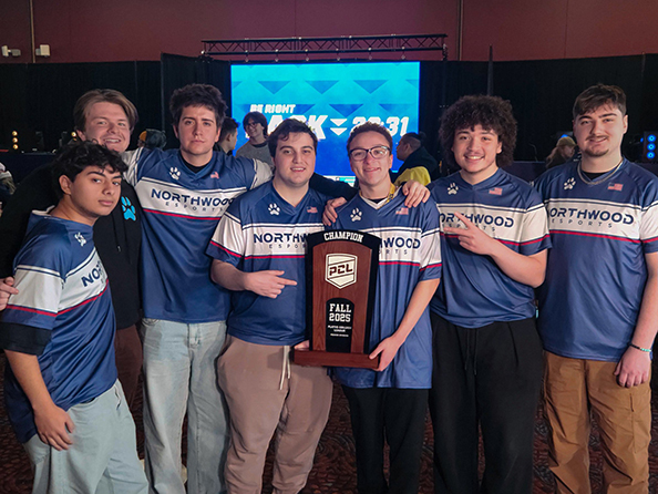 A group of seven young men in matching blue "Northwoods Esports" jerseys pose indoors, holding a large "Champion Fall 2023" trophy.