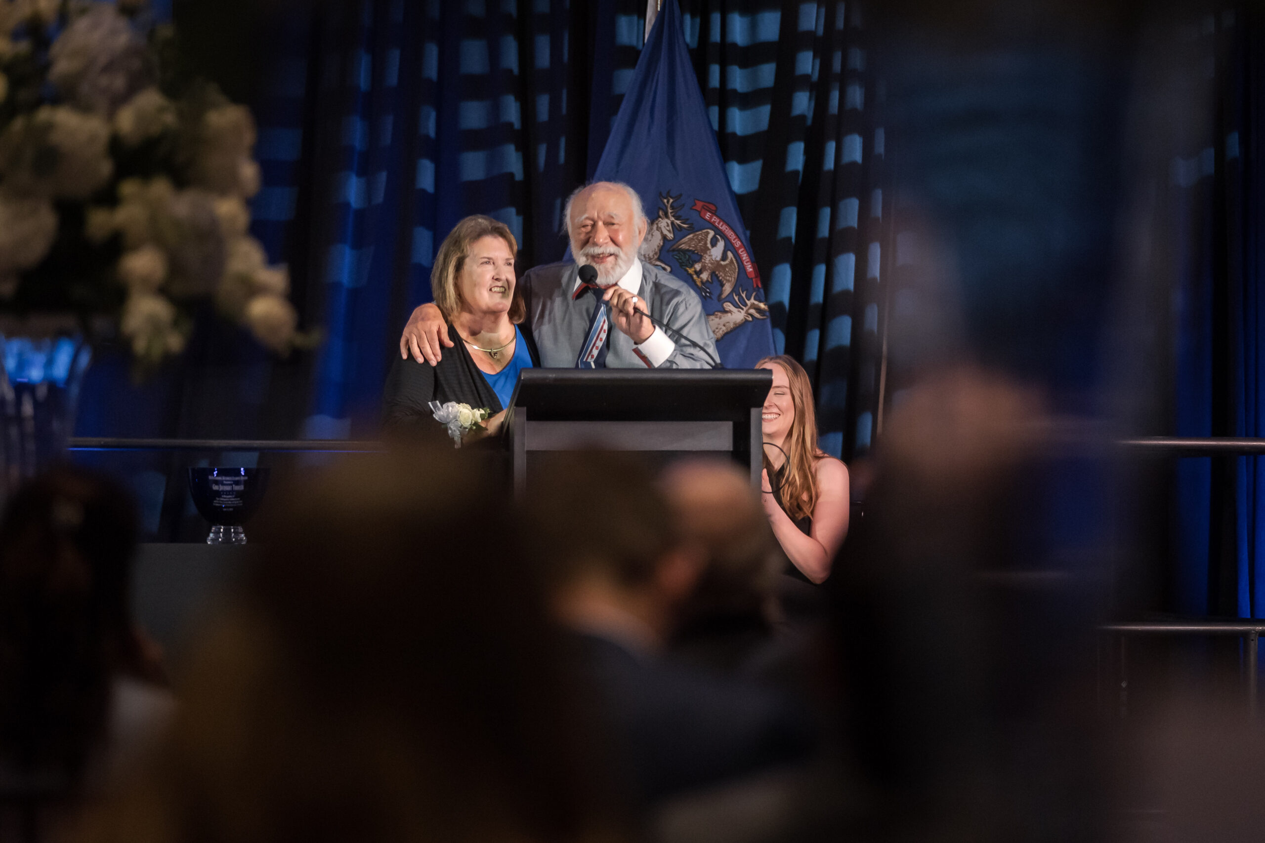 An older man and woman stand together at a podium, speaking to an audience in a formal setting with a state flag in the background.