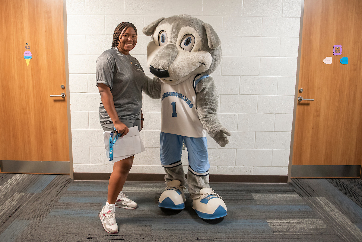 A smiling person stands next to a wolf mascot wearing a sports jersey and shorts in a hallway with closed wooden doors, celebrating the benefits enjoyed by Legacy students.