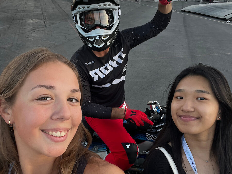 Two women take a selfie with a motocross rider in gear making a peace sign, set outdoors with event barriers and buildings—a lively scene capturing the spirit of automotive culture and mobility studies.