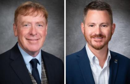 Two men in business attire pose against a dark, textured background; the man on the left has red hair and wears a suit with a plaid vest, while the man on the right has short brown hair and a blue blazer.