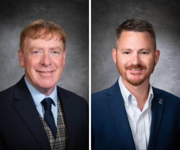 Two men in business attire pose against a dark, textured background; the man on the left has red hair and wears a suit with a plaid vest, while the man on the right has short brown hair and a blue blazer.