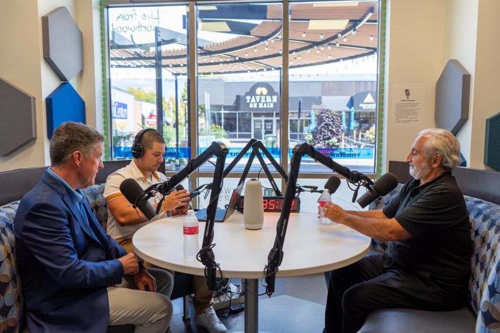 Three men sit around a round table with microphones, recording a podcast in a studio with a window showing a street scene outside.