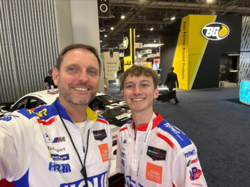 Two people in racing team uniforms smile for a selfie at an indoor automotive event, with race cars and promotional booths visible in the background—an exciting backdrop for those passionate about Automotive and Mobility Studies.