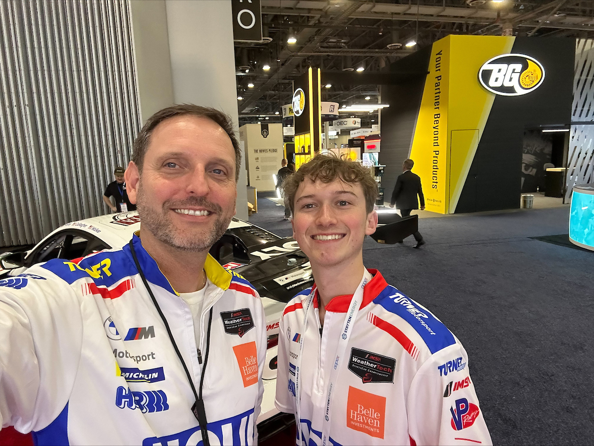 Two people in racing team uniforms smile for a selfie at an indoor automotive event, with race cars and promotional booths visible in the background—an exciting backdrop for those passionate about Automotive and Mobility Studies.