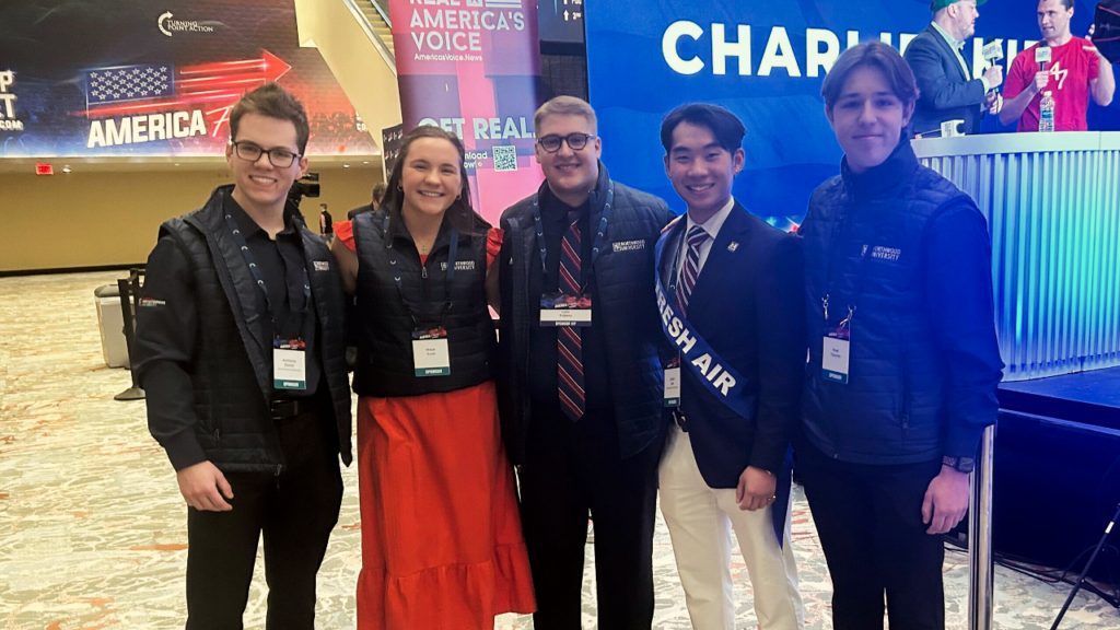 Five young adults wearing conference badges stand indoors at a Turning Point USA Northwood event, posing together and smiling in front of colorful banners and a stage.