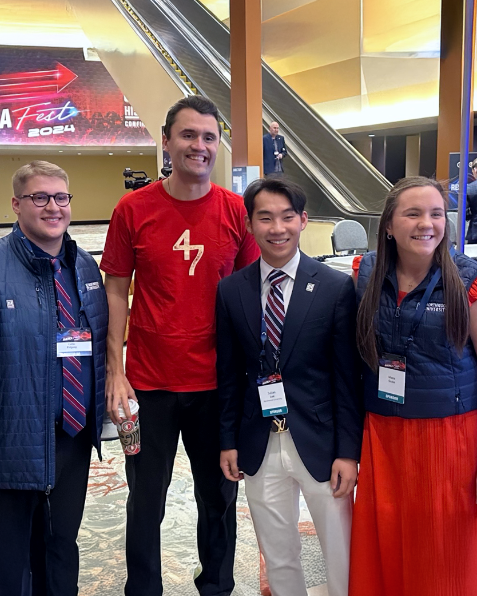 Four people pose together indoors at an event; three wear business attire, one sports a red "47" t-shirt.
