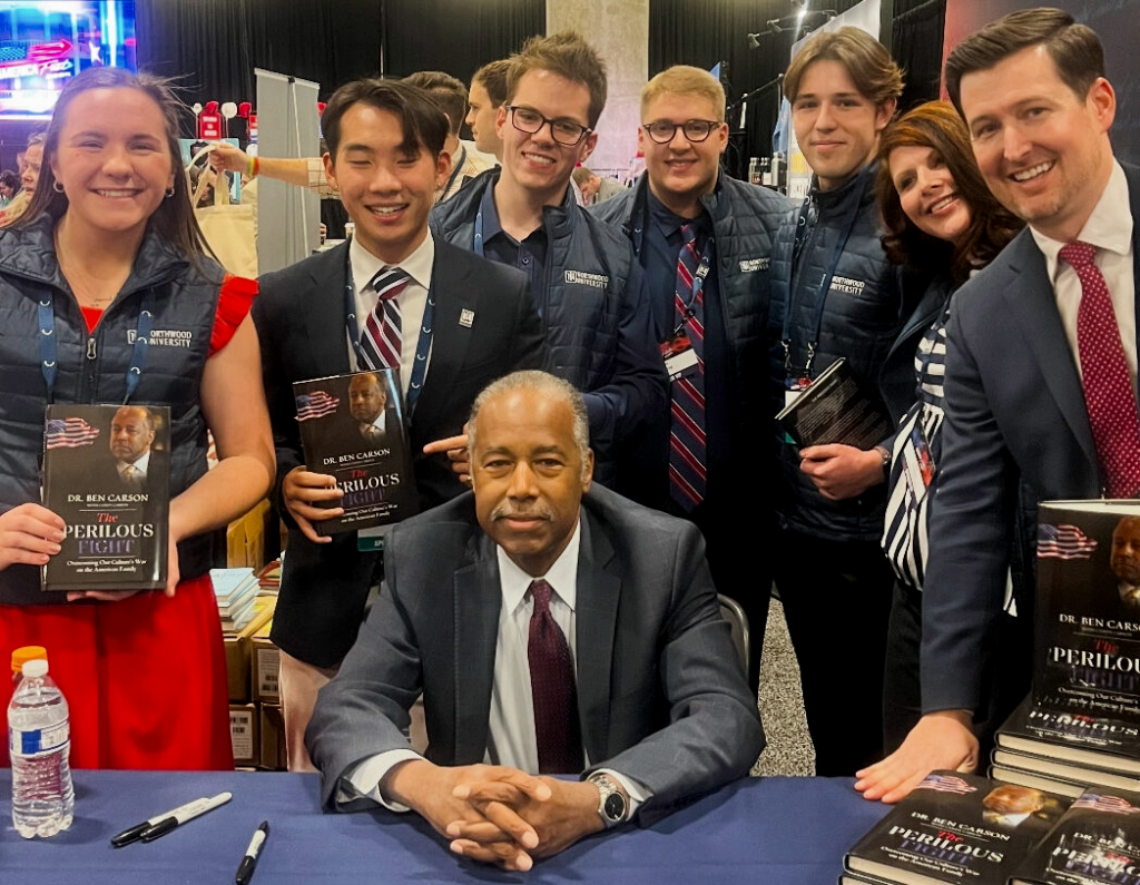 A group of young adults stands around a seated man at a Turning Point USA Northwood book signing event, holding copies of "Perilous Fight" as tables display stacks of the book.