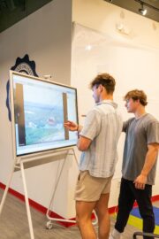 Two young men interact with a large touchscreen display showing an image of a cable car over a landscape in a brightly lit indoor space.