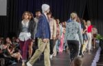 Models walk down a runway during a fashion show, wearing a variety of colorful and stylish outfits, with an audience seated on both sides of the stage.