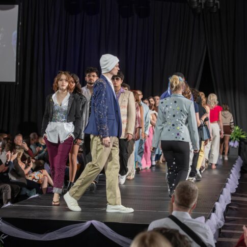 Models walk down a runway during a fashion show, wearing a variety of colorful and stylish outfits, with an audience seated on both sides of the stage.