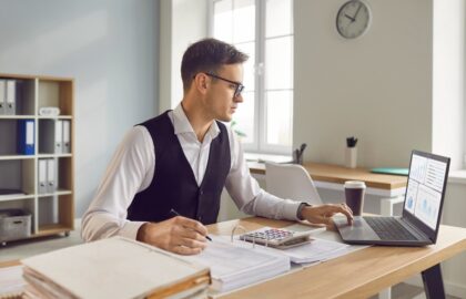 A man in business attire works at a desk on accounting tasks with a laptop, calculator, papers, and coffee cup in a bright office with shelves and a wall clock in the background.