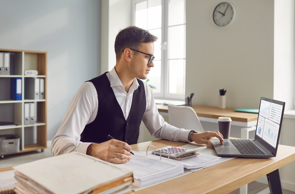 A man in business attire works at a desk on accounting tasks with a laptop, calculator, papers, and coffee cup in a bright office with shelves and a wall clock in the background.