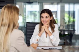 Two women sit across a desk in a modern office, engaged in conversation about Applied Management. One woman is smiling and listening, while the other holds a pen and papers.