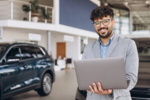 Man in a gray suit jacket holding a laptop, standing in a modern car showroom with vehicles in the background, showcasing expertise in Automotive Aftermarket Management.