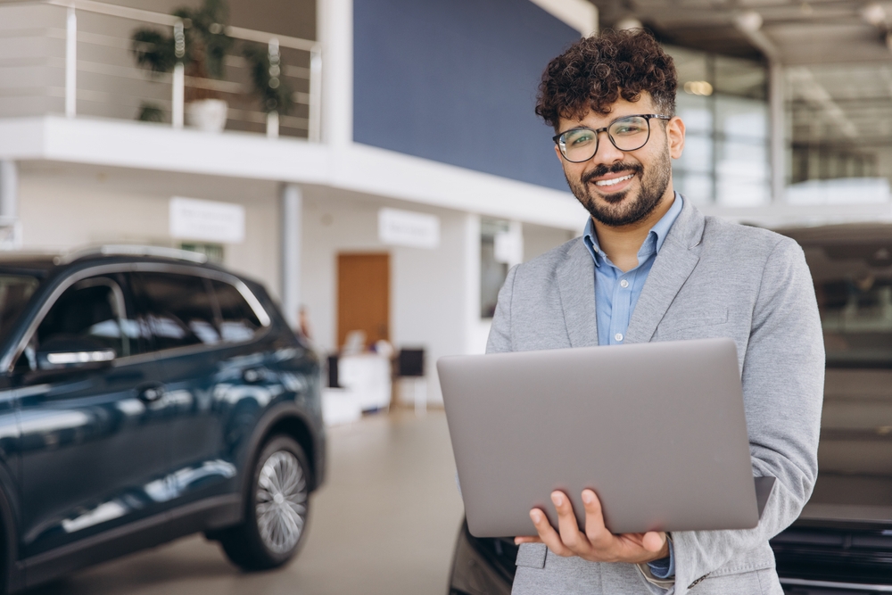 Man in a gray suit jacket holding a laptop, standing in a modern car showroom with vehicles in the background, showcasing expertise in Automotive Aftermarket Management.