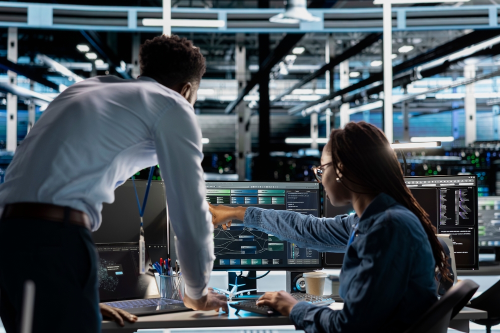 Two people in an office collaborate at a computer, applying their computer science expertise while analyzing data and pointing at information displayed on multiple monitors.
