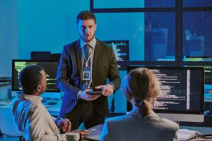 A man in a suit with a badge discusses cybersecurity management with two seated colleagues in an office, as computer monitors display code.