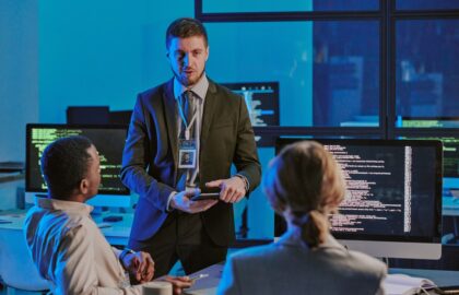A man in a suit with a badge discusses cybersecurity management with two seated colleagues in an office, as computer monitors display code.