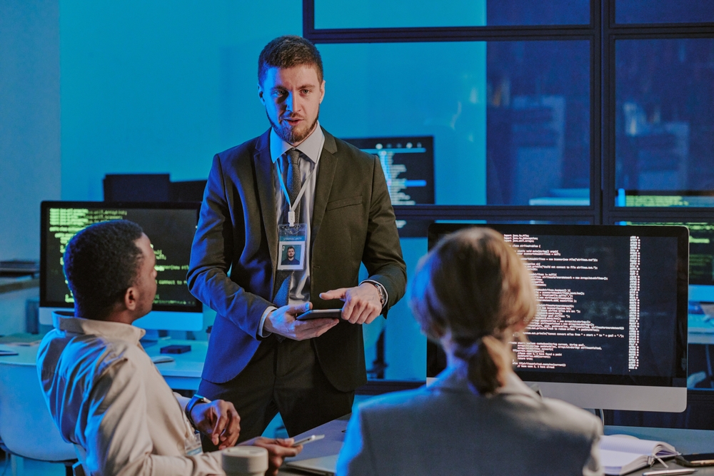 A man in a suit with a badge discusses cybersecurity management with two seated colleagues in an office, as computer monitors display code.