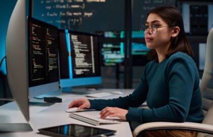 A person sits at a desk working on multiple computer monitors displaying code, with a tablet nearby and programming notes on a board in the background, immersed in Computer Science tasks.
