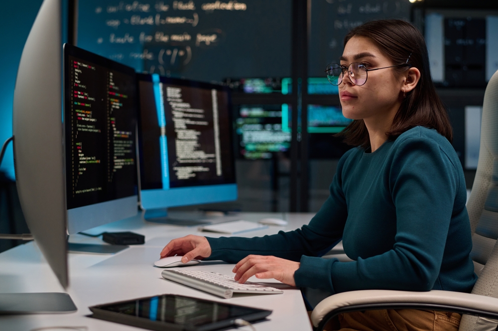 A person sits at a desk working on multiple computer monitors displaying code, with a tablet nearby and programming notes on a board in the background, immersed in Computer Science tasks.