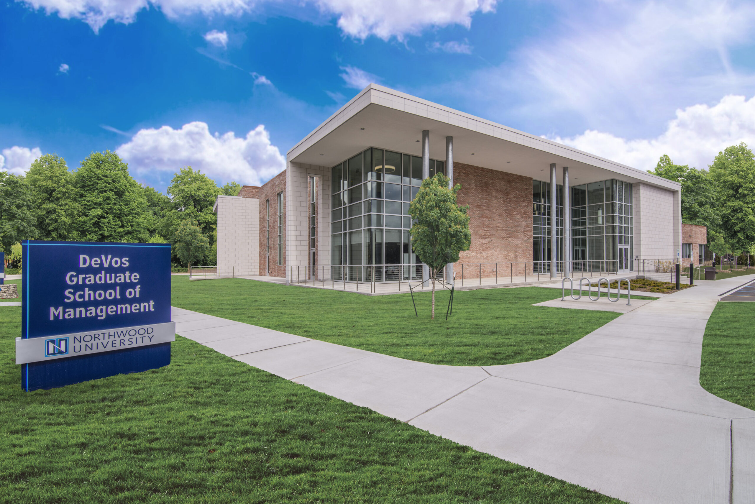 Modern building with large glass windows labeled DeVos Graduate School of Management, Northwood University, home to the Northwood University MBA in Health Leadership & Innovation, surrounded by green lawns and trees under a blue sky.