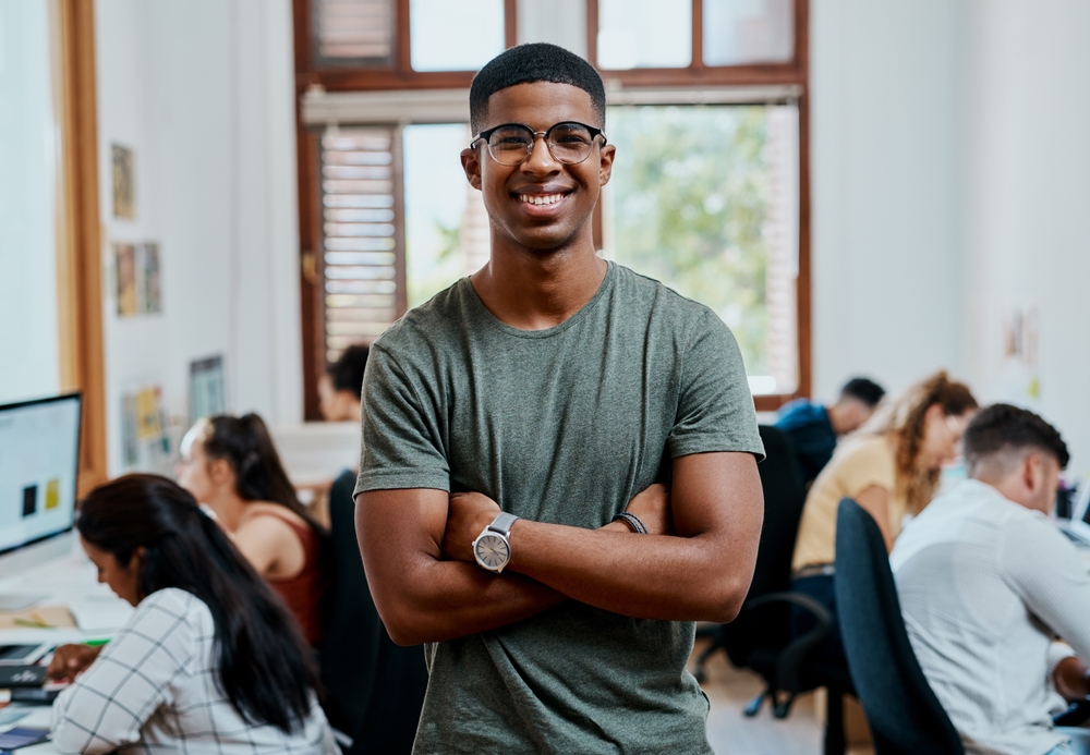 A man wearing glasses and a green t-shirt stands with arms crossed, smiling in an office with colleagues working at desks in the background.