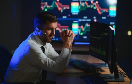 A man wearing a headset sits at a desk, analyzing accounting data and financial charts on his monitors in a dark office with large screens in the background.
